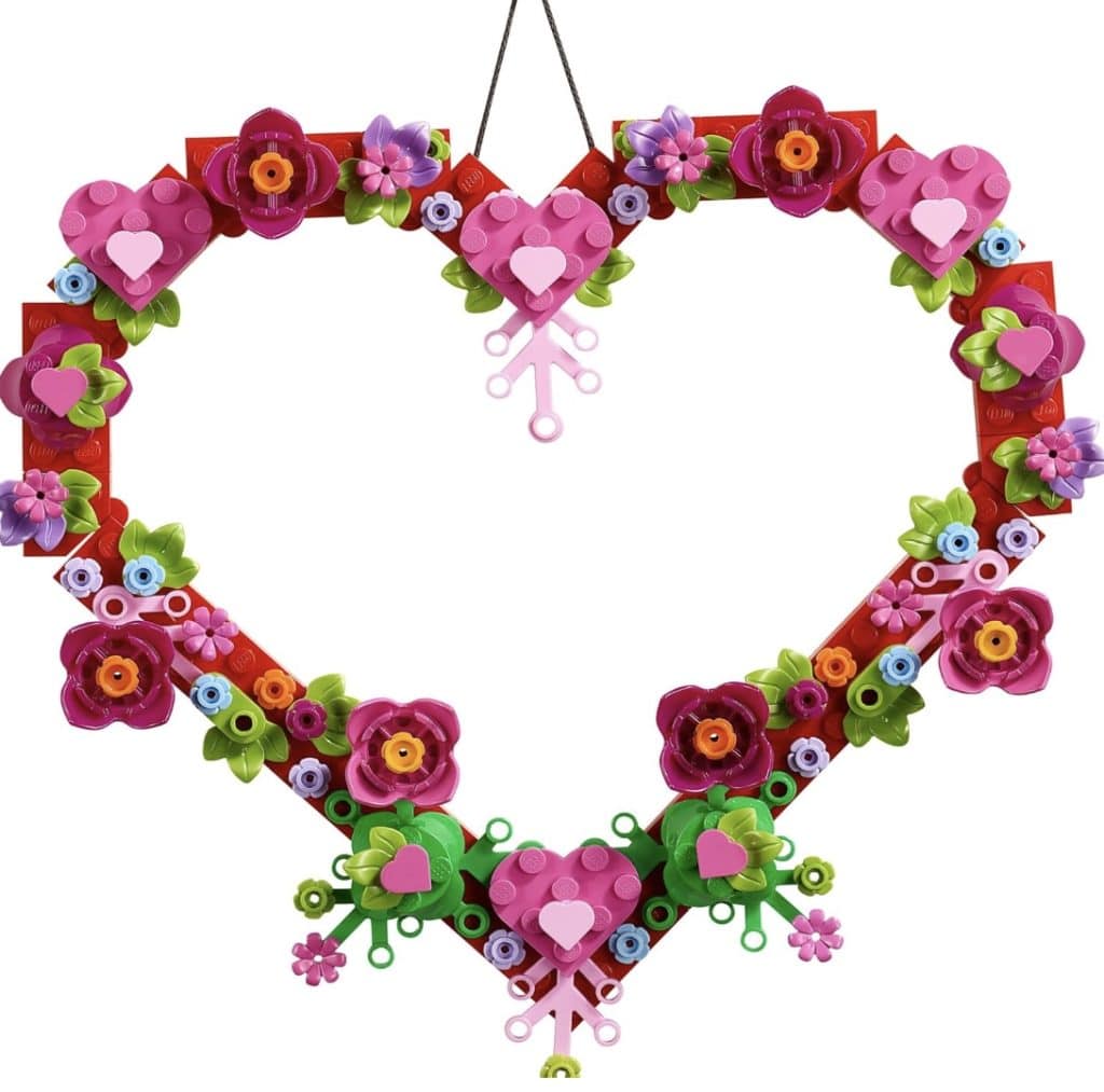 A heart-shaped seasonal wreath crafted from colorful LEGO pieces, featuring pink and red flowers, green leaves, and small decorative elements. This symmetrical design hangs gracefully from a black string.